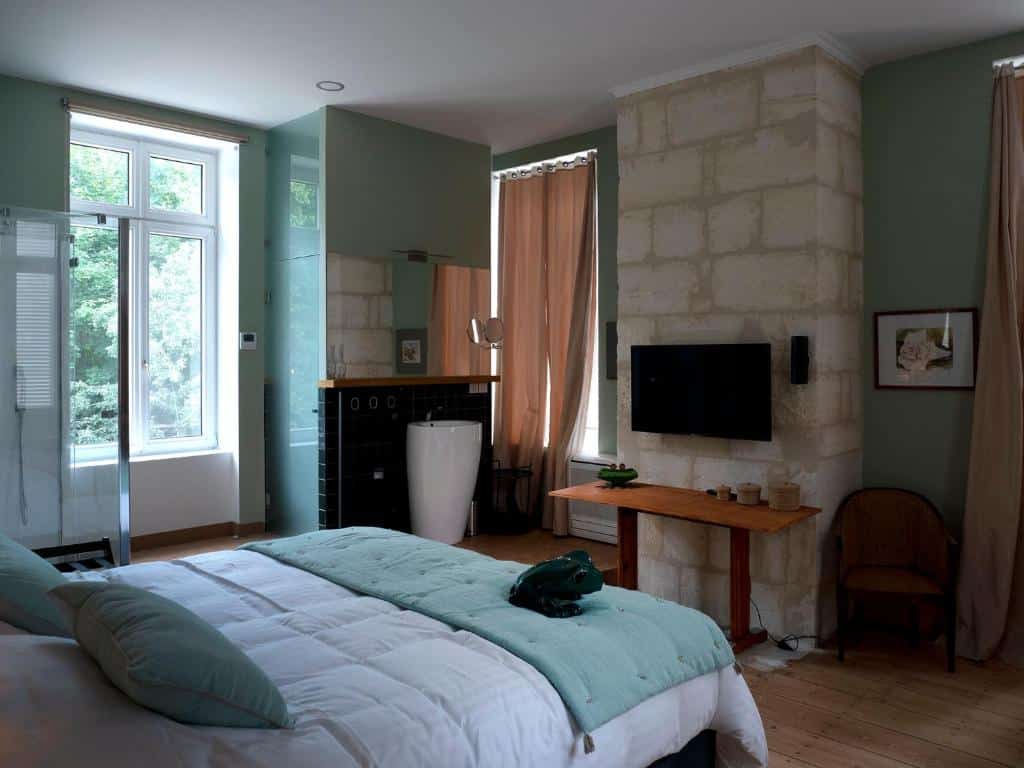 Chambre d'hôtel moderne aux murs vert et pierre. Grand lit blanc, salle de bain ouverte avec douche et lavabo. Fenêtre lumineuse, TV, table en bois.
