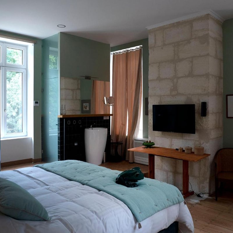 Chambre d'hôtel moderne avec lit blanc et jeté vert, salle de bain ouverte avec lavabo design, mur en pierre, et statue de grenouille.