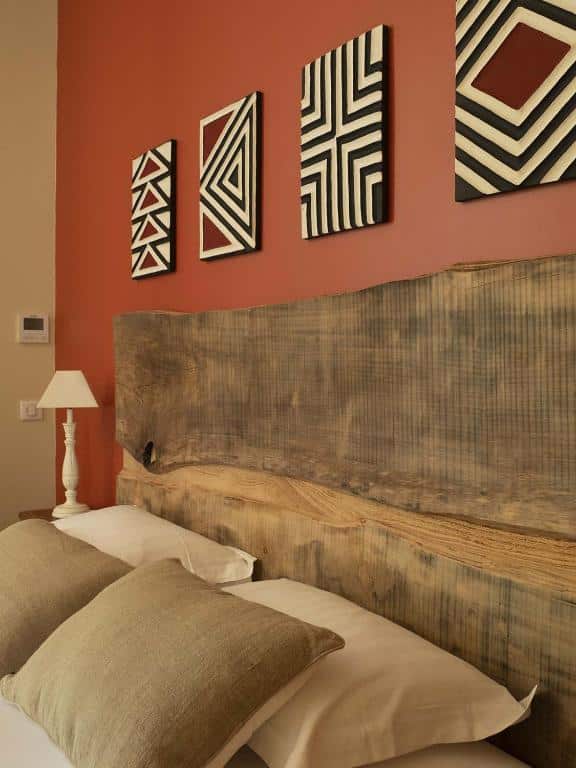 Chambre avec tête de lit en bois brut. Mur terracotta orné de quatre tableaux géométriques noirs et blancs. Lit avec coussins beiges.