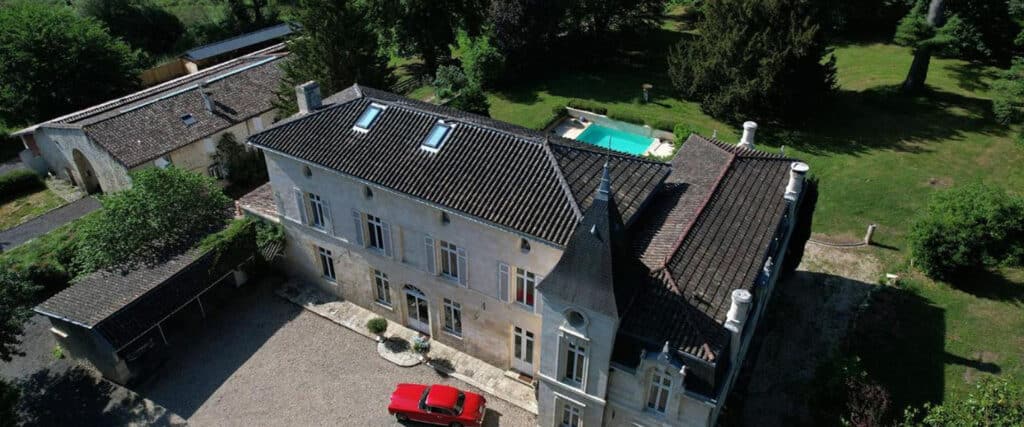 Vue aérienne d'un grand château en pierre avec toits sombres, tourelle, piscine turquoise et voiture rouge garée.