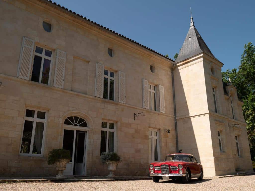 Élégant château en pierre avec tour et fenêtres à volets blancs, devant lequel est garée une voiture classique rouge sur gravier.