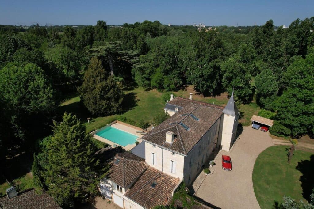 Domaine de caractère avec grande maison en pierre, tour, piscine bleue et vaste parc arboré. Deux voitures classiques sur la cour.