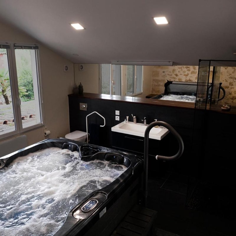 Jacuzzi intérieur bouillonnant dans une suite moderne avec lavabo blanc, WC et grandes fenêtres. Espace détente noir et bois.