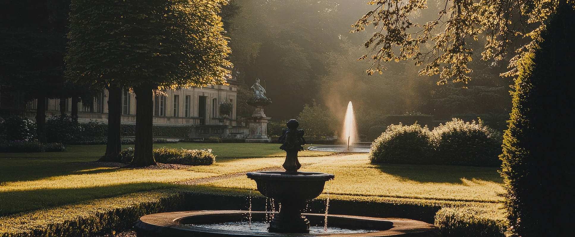 Jardin historique avec fontaines et grand bâtiment. Rayons de soleil dorés à travers la brume du matin sur l'herbe et les arbres.