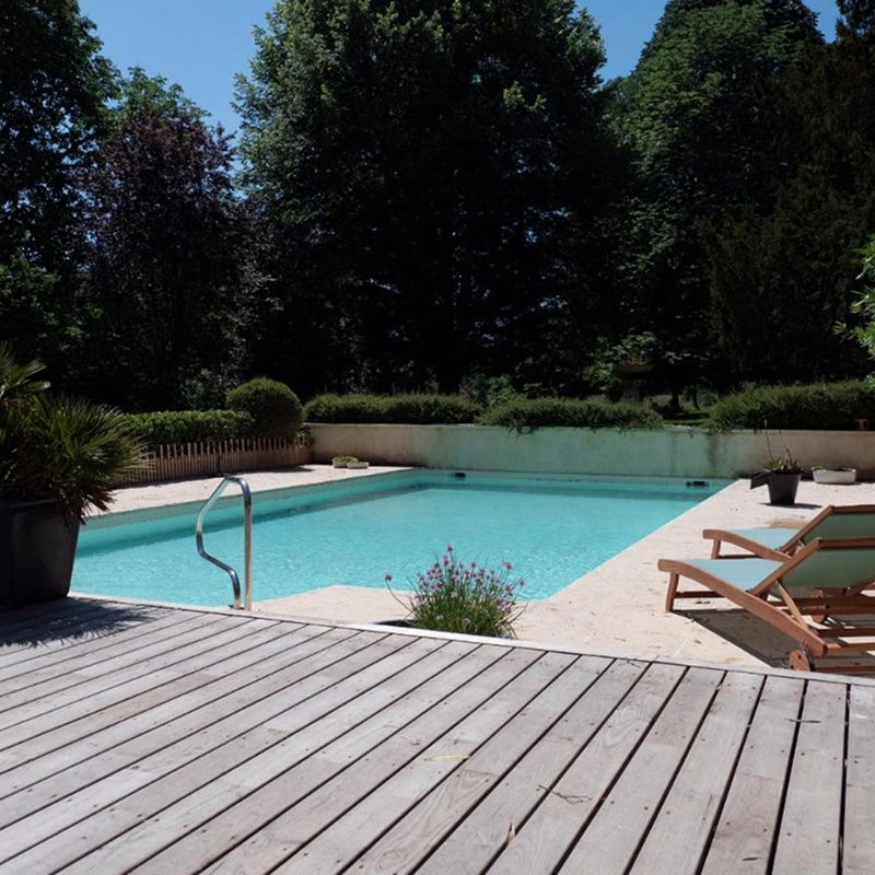 Piscine extérieure bleue avec terrasse en bois claire, transats et végétation luxuriante. Lumière du soleil sur l'eau.