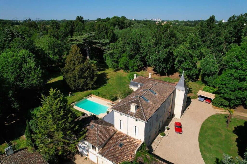 Vue aérienne d'une grande propriété avec maison élégante, tour, piscine turquoise, vaste parc arboré et voitures classiques.