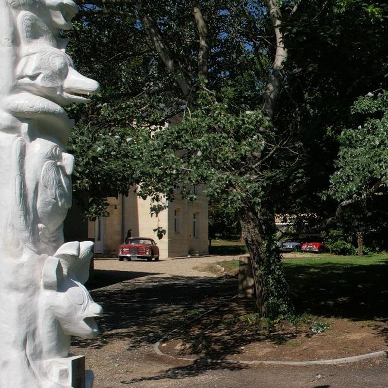Sculpture blanche d'animaux, château avec voiture ancienne rouge sur allée gravillonnée, et deux voitures classiques dans un parc boisé.
